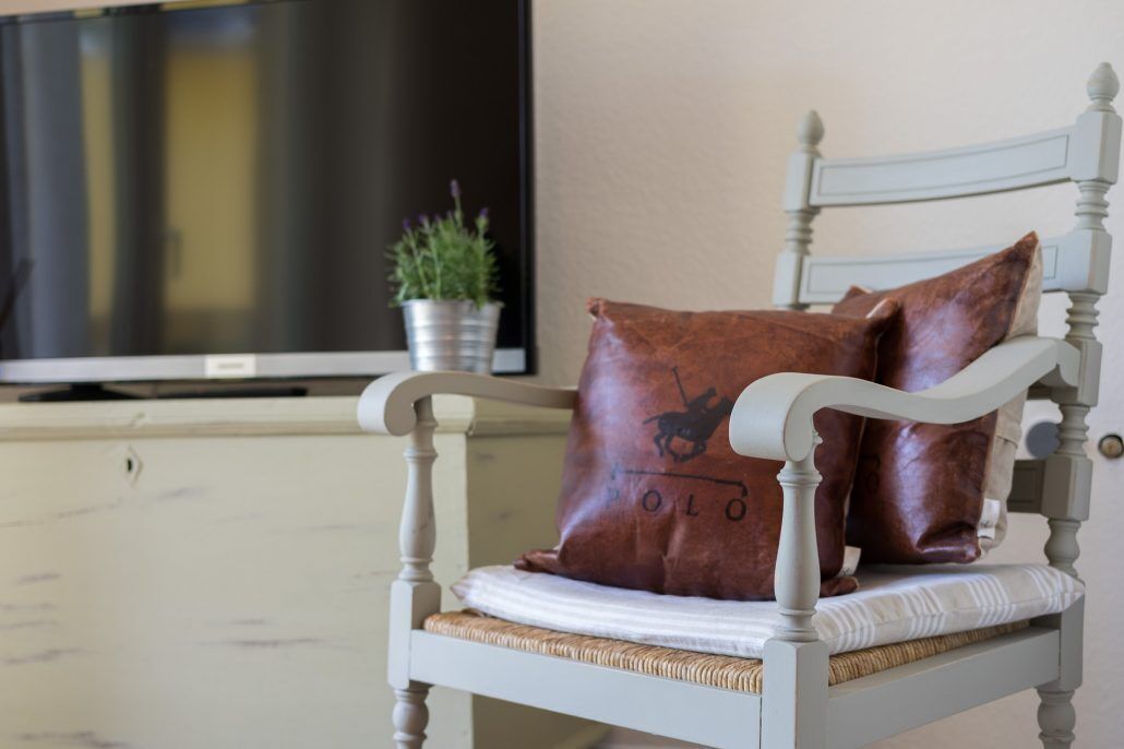 A white wooden chair with a woven seat cushion, a brown leather pillow with a horse logo, and a television on a stand in the background.