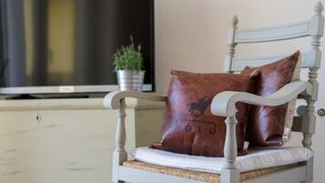 A white wooden chair with a woven seat cushion, a brown leather pillow with a horse logo, and a television on a stand in the background.