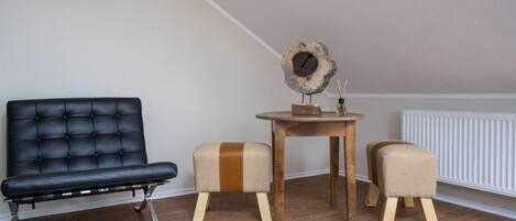 Un salon moderne avec un canapé en cuir noir, deux tabourets en bois, une table ronde en bois sur laquelle se trouve une horloge, et un tapis en fourrure sur le sol.