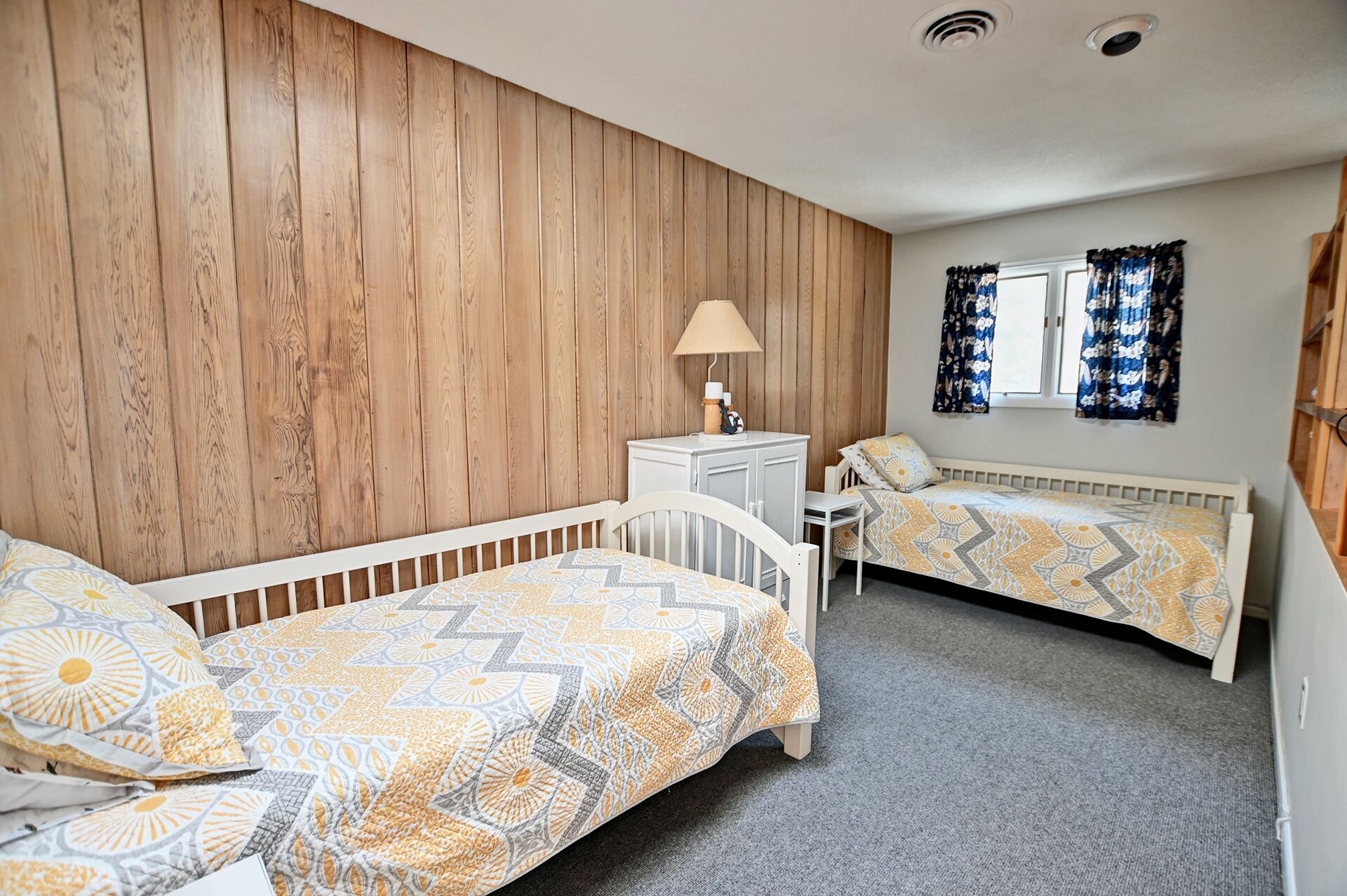 A bedroom with two single beds, a small white dresser, a lamp, and a window with curtains.