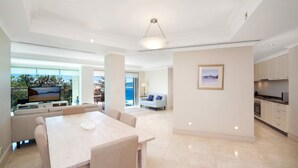 A spacious living area with a dining table, sofas, and a TV displaying a beach scene. - Unit 14  - 3 Bed Garden View (TERRIGAL)