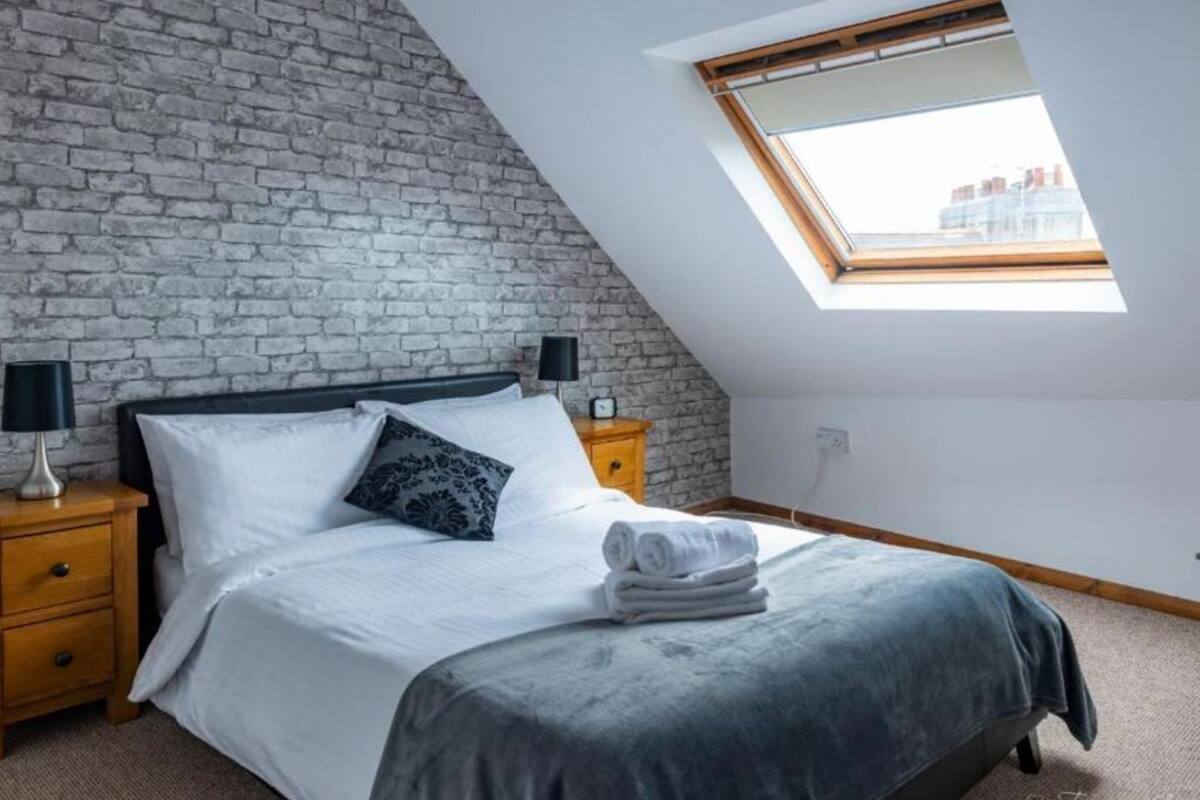 A bedroom with a sloped ceiling, a skylight, a bed with white linens, a grey blanket, and a brick wall.