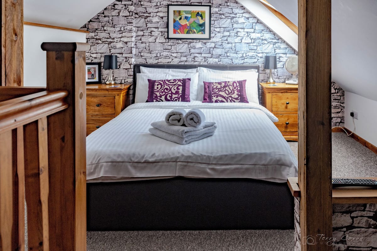 A bedroom with a stone wall, a bed with white and purple bedding, two bedside tables with lamps, and a framed picture on the wall.