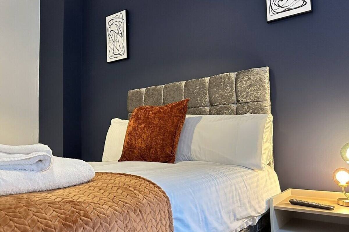 A neatly made bed with a brown quilt, white linens, and a stone-patterned headboard. There is a remote control on a bedside table with a lamp.