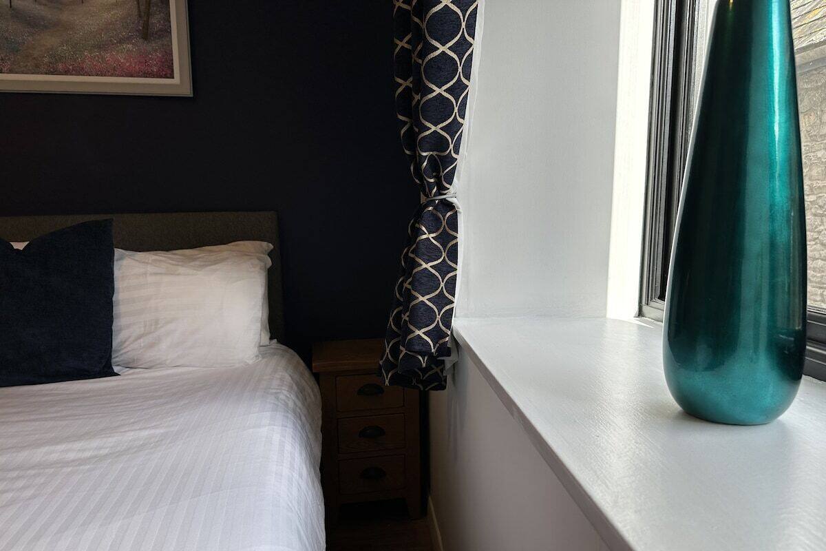 A bedroom with a bed, a nightstand, a window with curtains, and a vase on the windowsill.