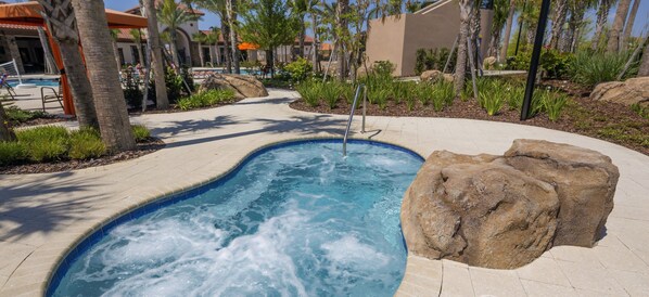A hot tub with a rock on one side, surrounded by a landscaped garden with palm trees and a pool area in the background.