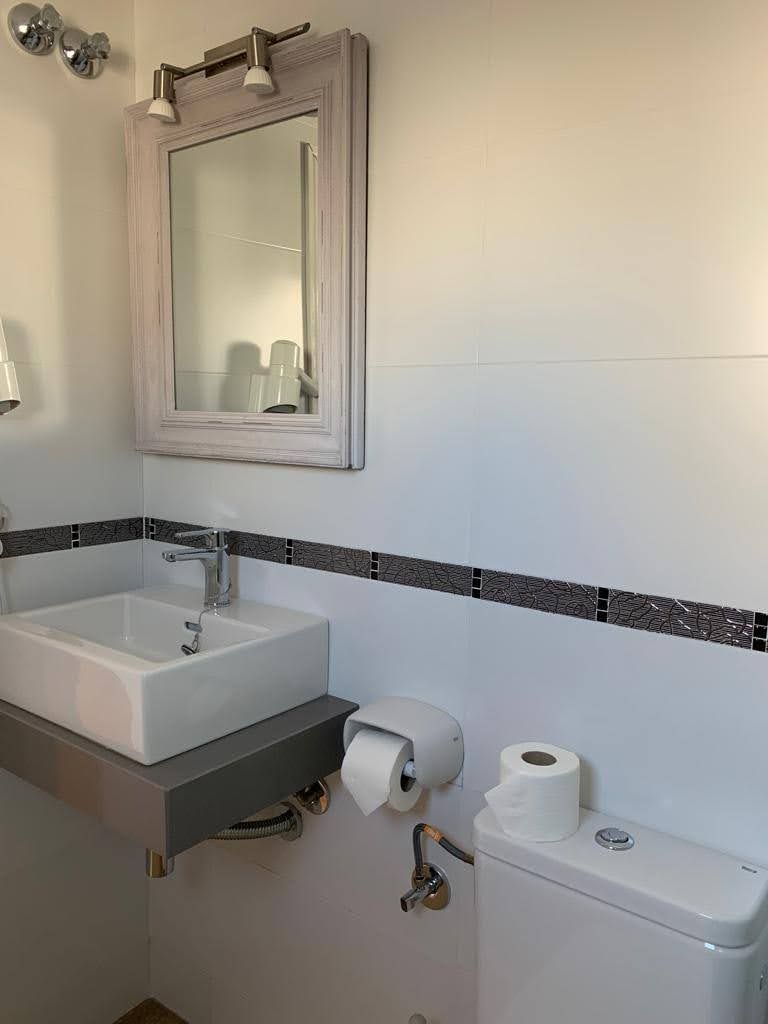 Un baño con un lavamanos blanco, un espejo encima y un inodoro con un rollo de papel higiénico a la derecha.