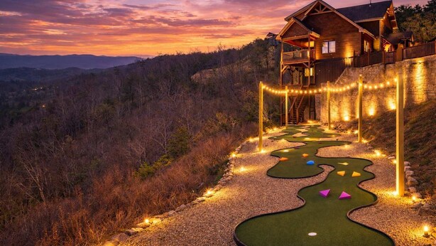 A wooden cabin with a deck, surrounded by a golf putting green with a winding path, set against a sunset backdrop. - Misty Bluff - Indoor pool & mountain views (Sevierville)