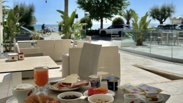 Preparación para el desayuno con cruasanes, bagels y un bol de fruta sobre una mesa bajo una sombrilla, con vista a una piscina y palmeras.