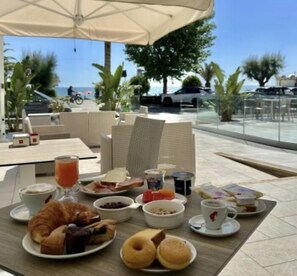 A breakfast setup with croissants, bagels, and a fruit bowl on a table under an umbrella, overlooking a pool and palm trees.