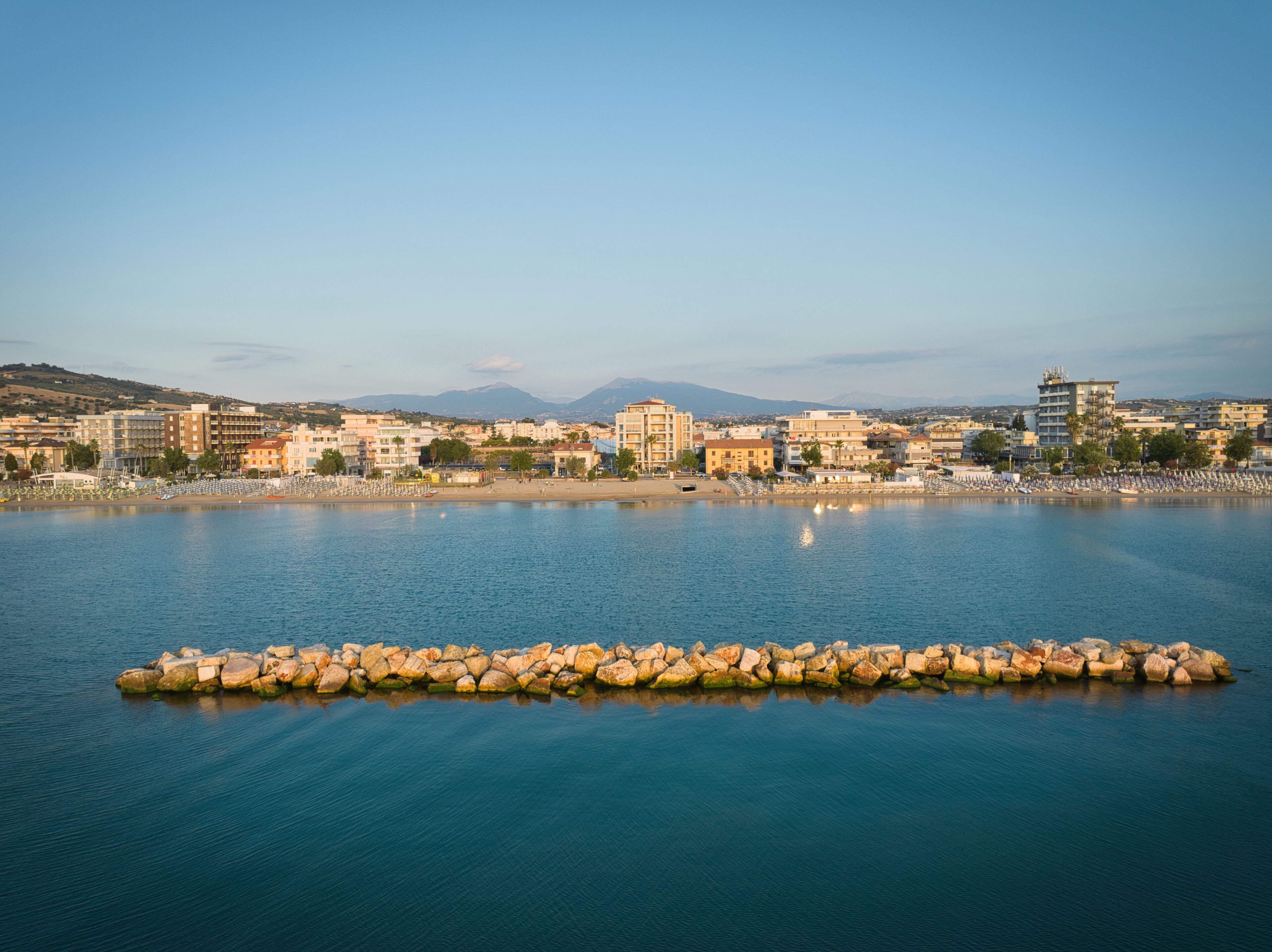 A coastal cityscape with a calm sea, buildings, and a rocky breakwater.