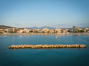 A coastal cityscape with a calm sea, buildings, and a rocky breakwater.