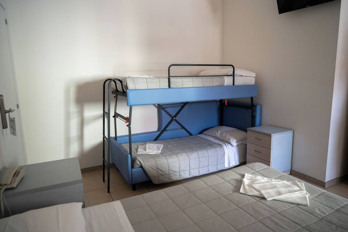 Una cama individual superpuesta con escritorio y un mueble pequeño, un teléfono sobre una mesa y una ventana con persianas.