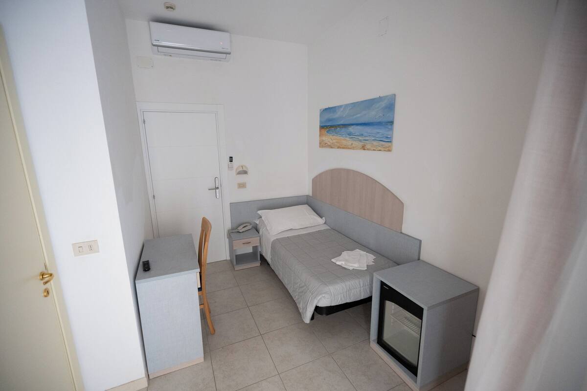 Una habitación pequeña y bien iluminada con una cama, un escritorio, una silla, un televisor y un cuadro en la pared.