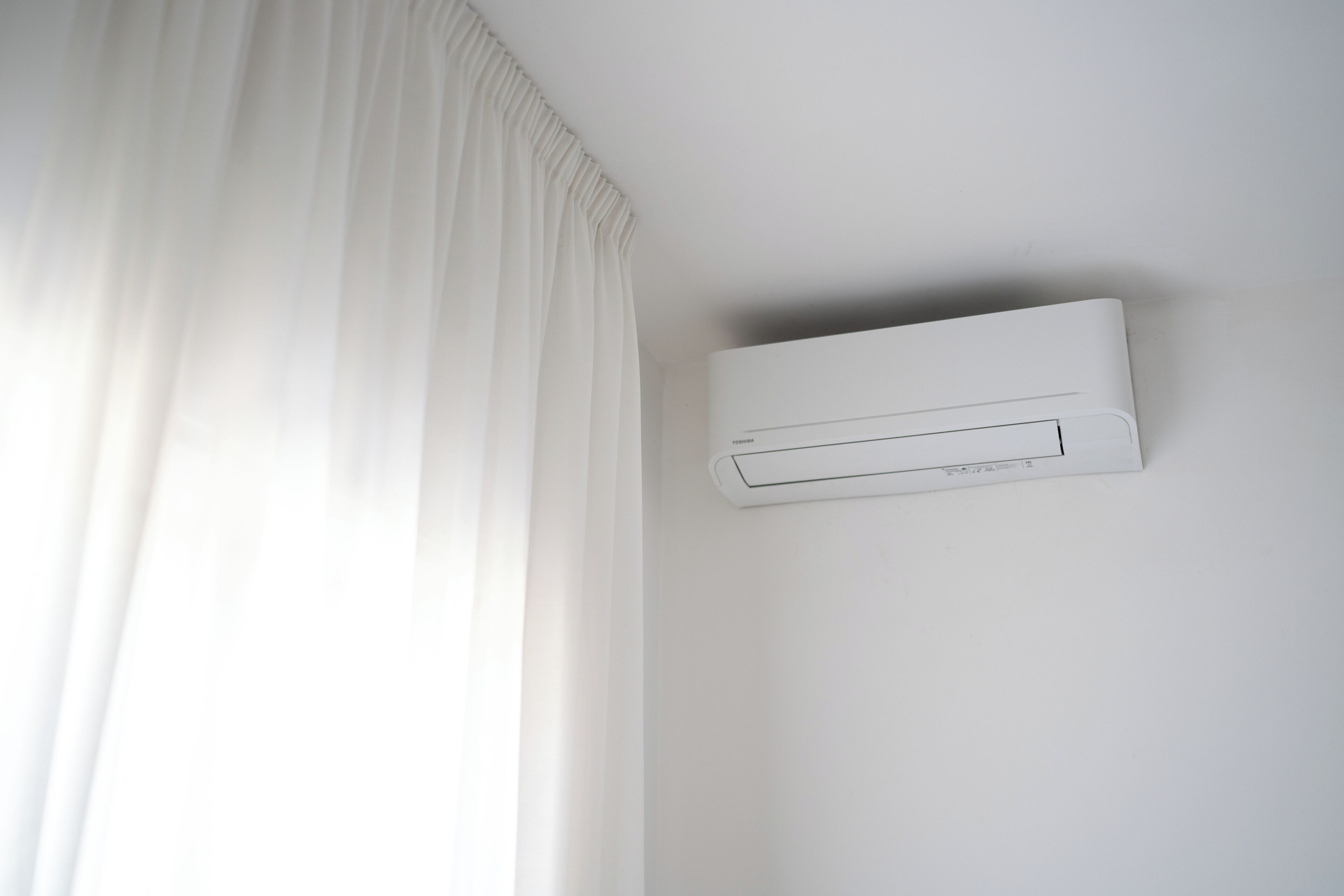 En hvid airconditionenhed monteret på en væg i et rum med hvide vægge og et hvidt gardin.
