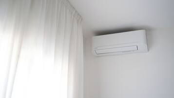 A white air conditioning unit mounted on a wall in a room with white walls and a white curtain.