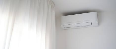 En hvid airconditionenhed monteret på en væg i et rum med hvide vægge og et hvidt gardin.