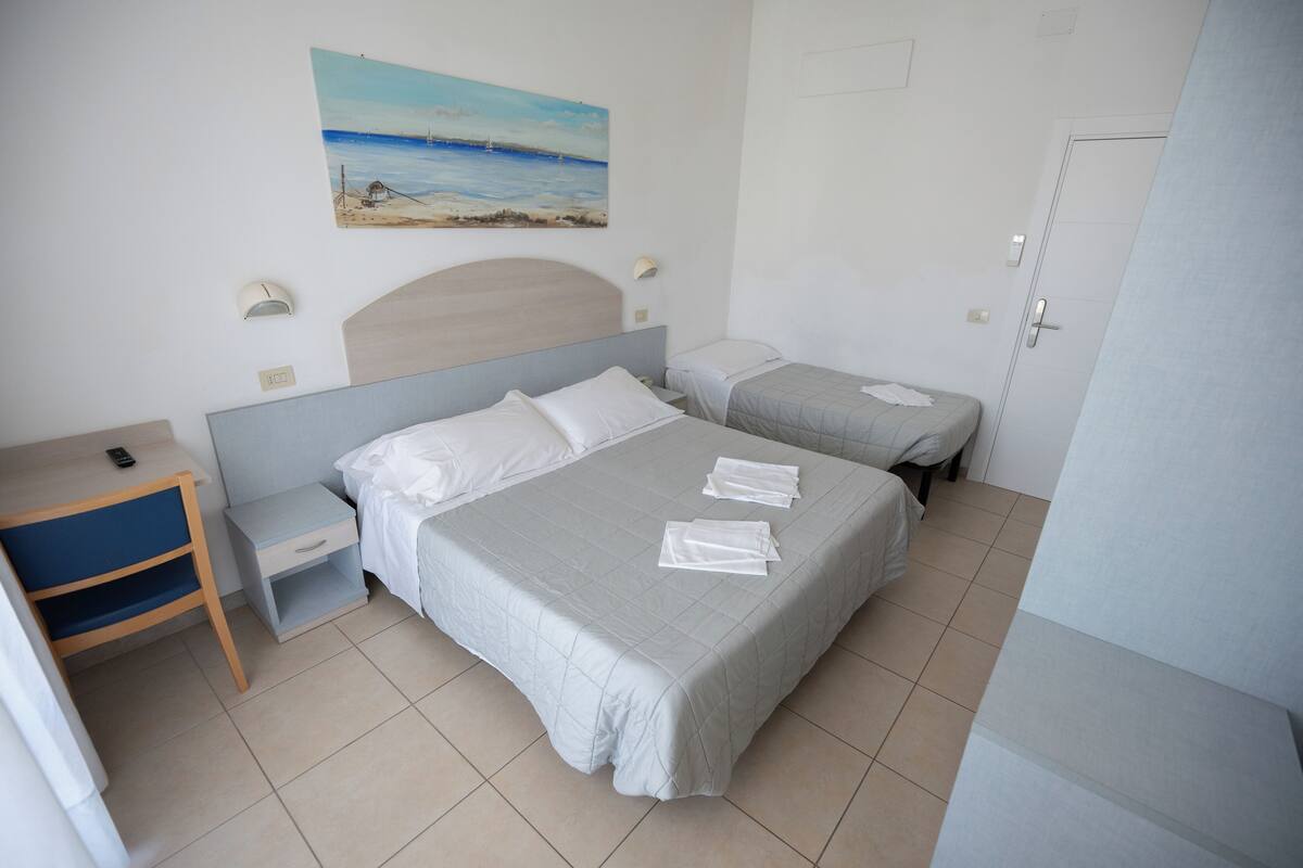Habitación de hotel con dos camas, un escritorio, una silla, un cuadro de playa y una puerta.