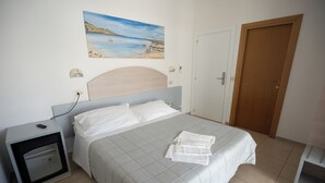 Una cama bien hecha con una colcha blanca y almohadas, una mesita de noche con una lámpara, un cuadro con una escena de playa en la pared y una puerta de madera.