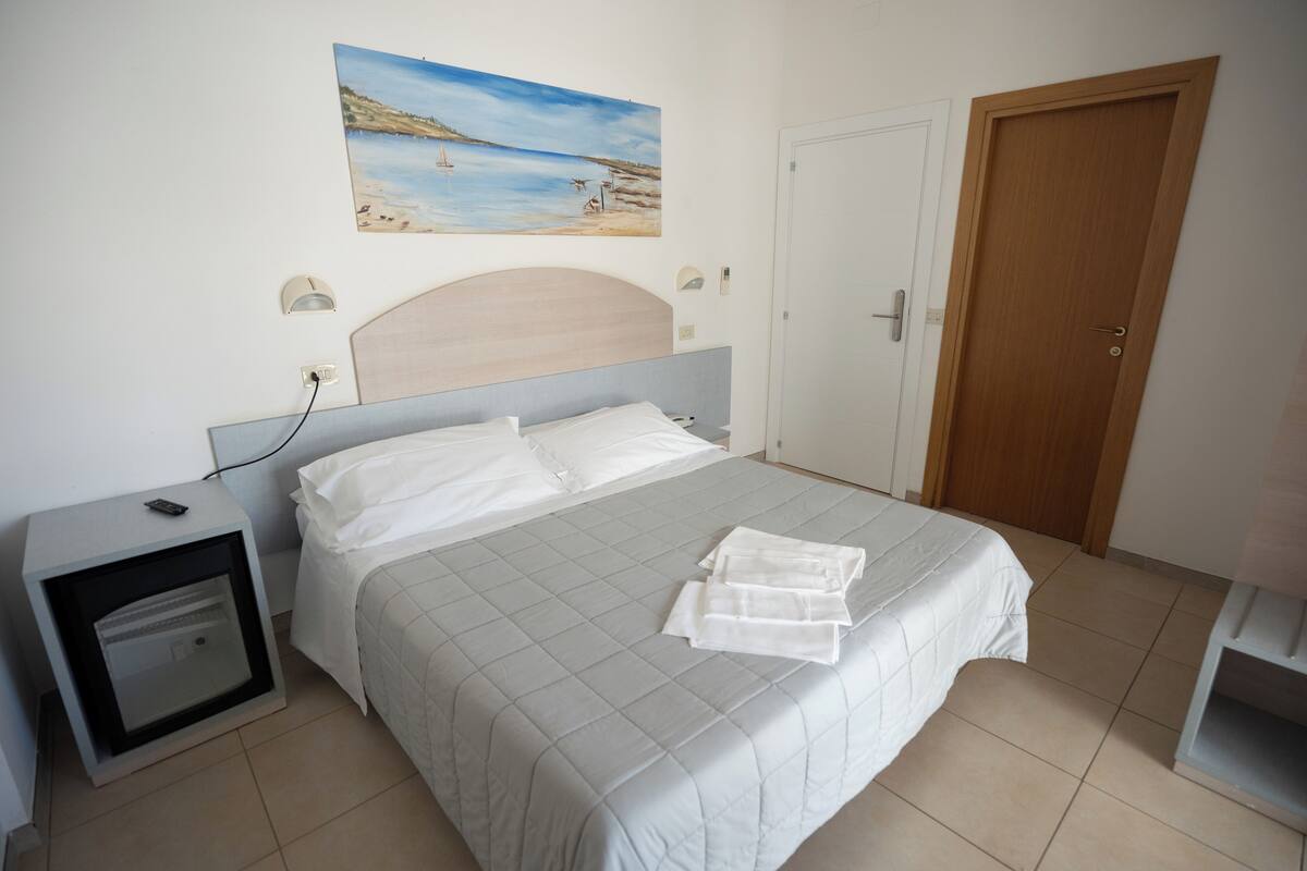 Una cama bien hecha con una colcha blanca y almohadas, una mesita de noche con una lámpara, un cuadro con una escena de playa en la pared y una puerta de madera.
