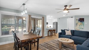Una sala de estar moderna con un sofá azul, una mesa de comedor de madera con sillas, una cocina al fondo y un ventilador de techo.