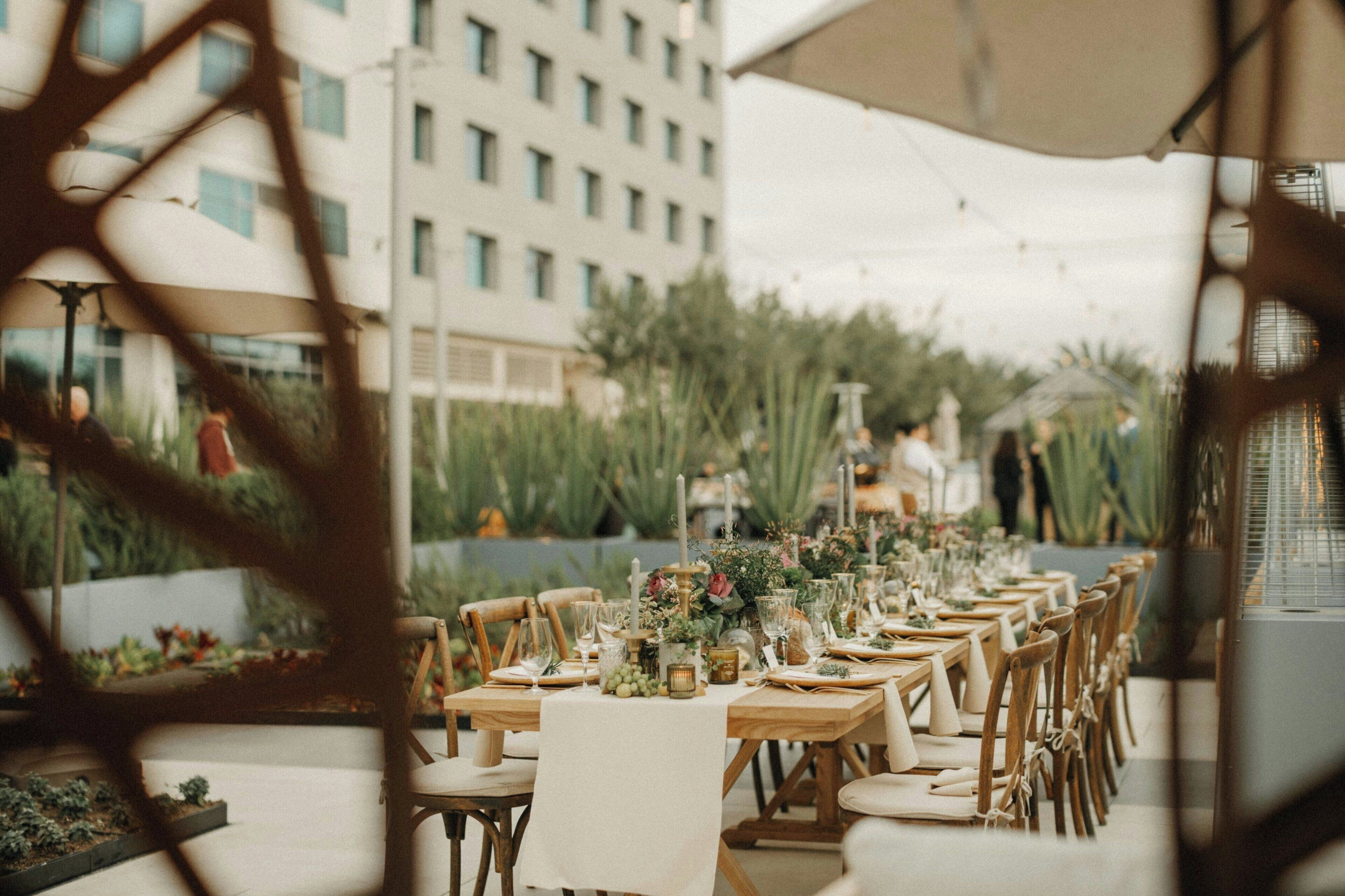 An outdoor dining area with long wooden tables set up for a meal, surrounded by greenery and a building in the background.