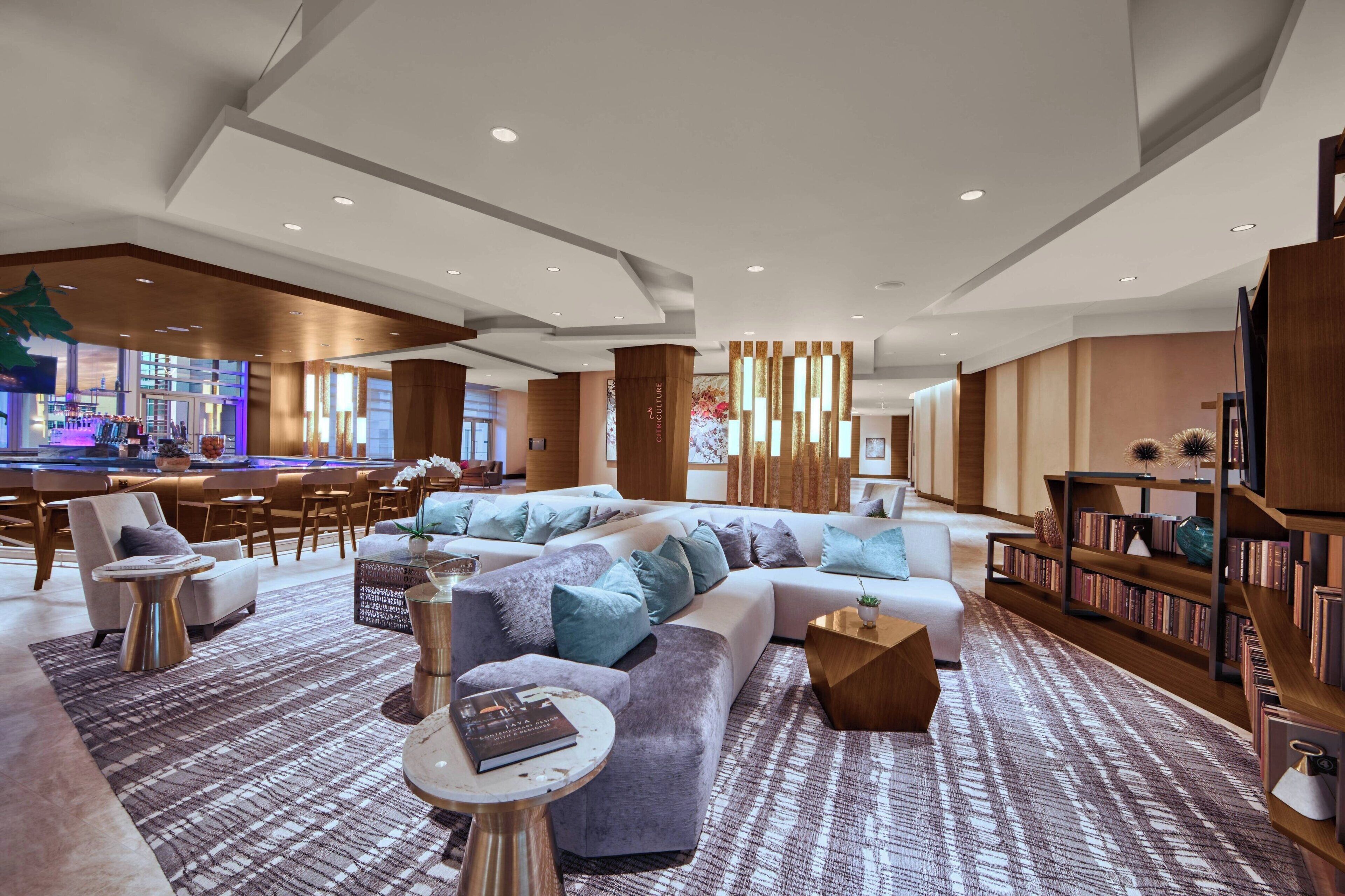 A modern hotel lobby with a large seating area, a bar, and a bookshelf.
