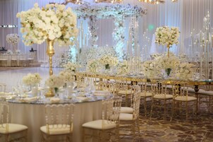 Uma sala de banquetes com mesas redondas, cadeiras douradas e centros de mesa florais brancos.