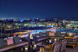 Terraza en lo alto con vista a una piscina, asientos exteriores y luces de la ciudad al fondo.