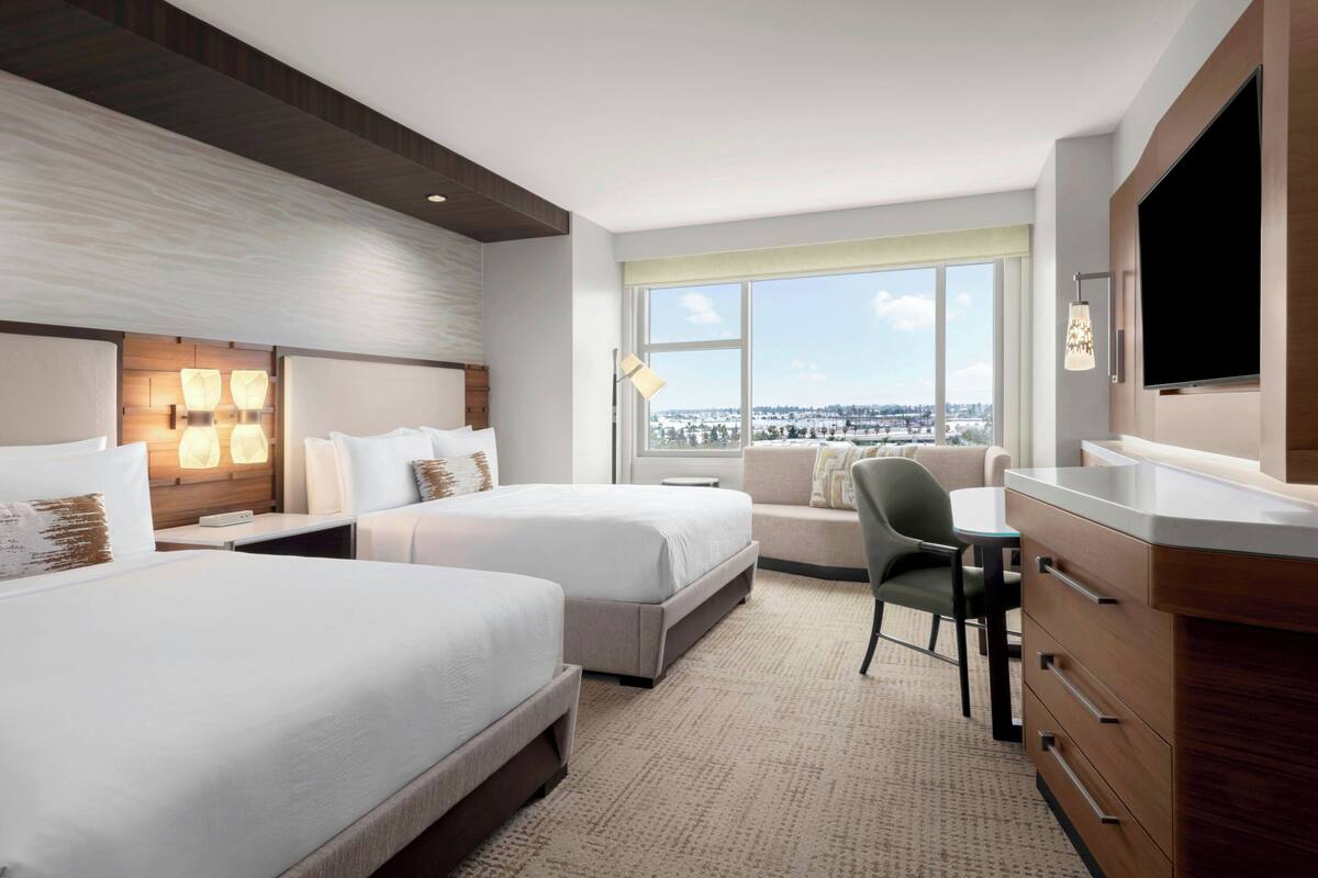A hotel room with two beds, a desk, a chair, a TV, and a large window offering a city view.