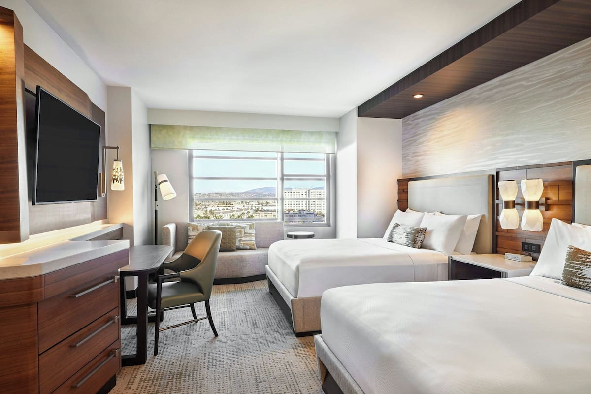 A hotel room with two beds, a desk, a chair, a TV, and a large window offering a city view.
