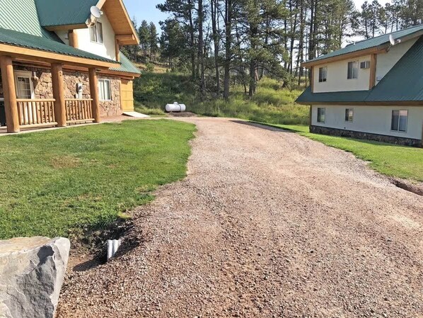 A two-story building with a stone foundation and a green roof, surrounded by a gravel driveway and a grassy area. - Cottage at Storm Mountain #5 (Hill City)