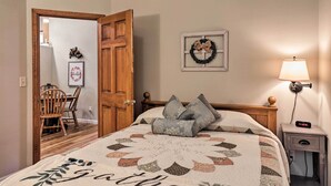 Un dormitorio con cama, una mesita de noche con lámpara, una puerta de madera, un cuadro enmarco y una pequeña mesa con sillas.