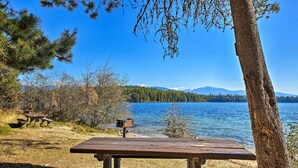 Uma área de piquenique à beira do lago, com um banco e uma mesa para piquenique, rodeada por árvores e uma vista deslumbrante do lago e montanhas ao longe.