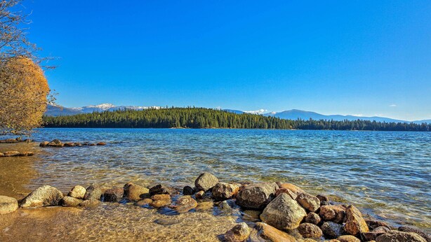 Um lago límpido com rochas visíveis em primeiro plano e uma cadeia montanhosa arborizada ao fundo.
