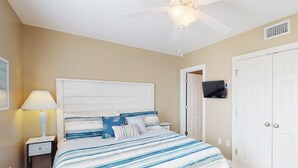 Une chambre à coucher comprenant un lit, une table de chevet, une lampe, un ventilateur de plafond, un téléviseur et une porte double.