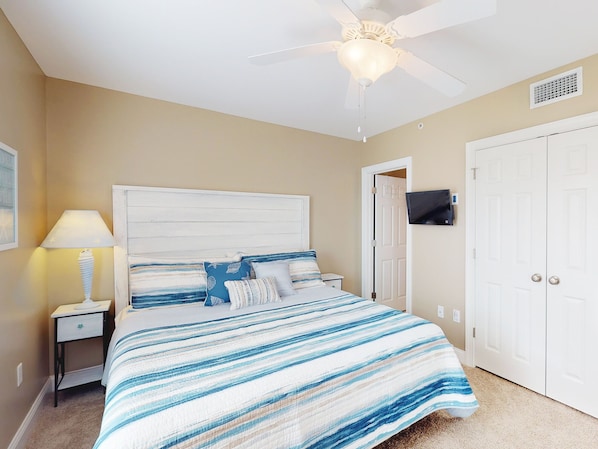 A bedroom with a bed, nightstand, lamp, ceiling fan, TV, and double doors. - Heated Pool Access | 5*Clean | Flex Cancelation (Tybee Island)
