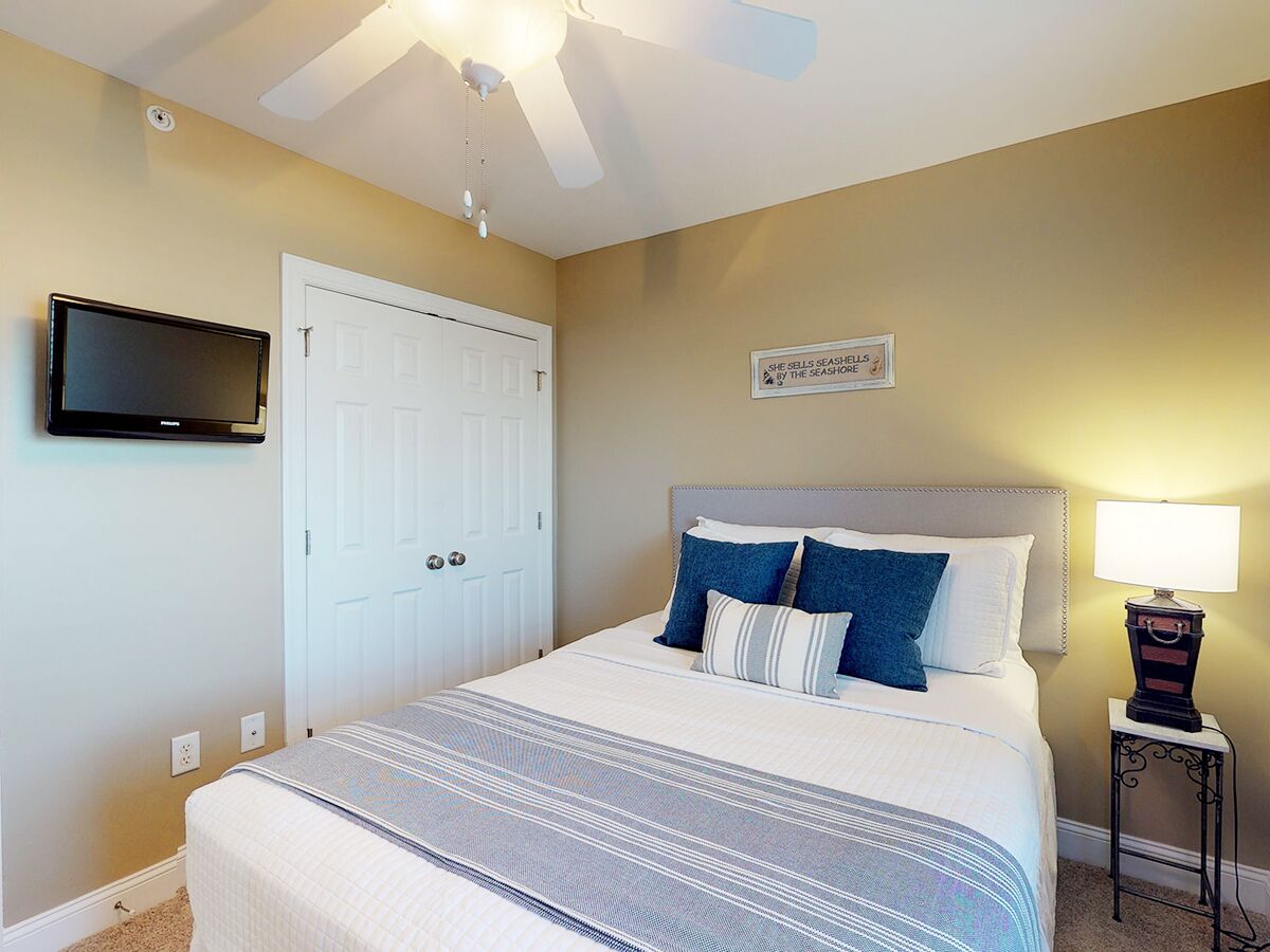 A bedroom with a bed, a TV mounted on the wall, a ceiling fan, and a bedside table with a lamp.