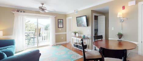 A living room with a ceiling fan, a TV mounted on the wall, a dining table with chairs, a sofa, and a large window with curtains.