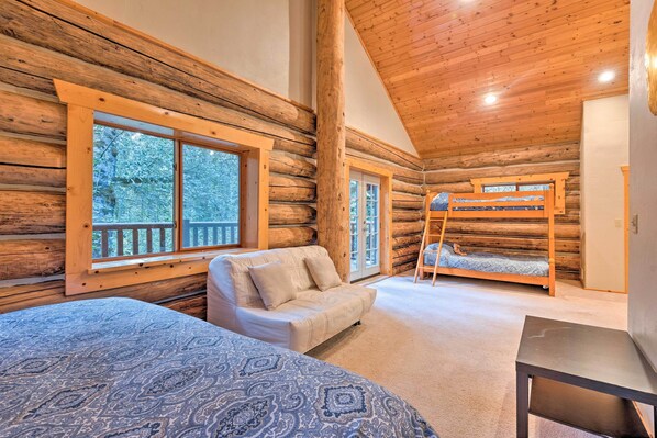 Une chambre rustique aménagée dans un chalet en rondins, avec un lit superposé, un canapé et une grande fenêtre donnant sur la forêt.