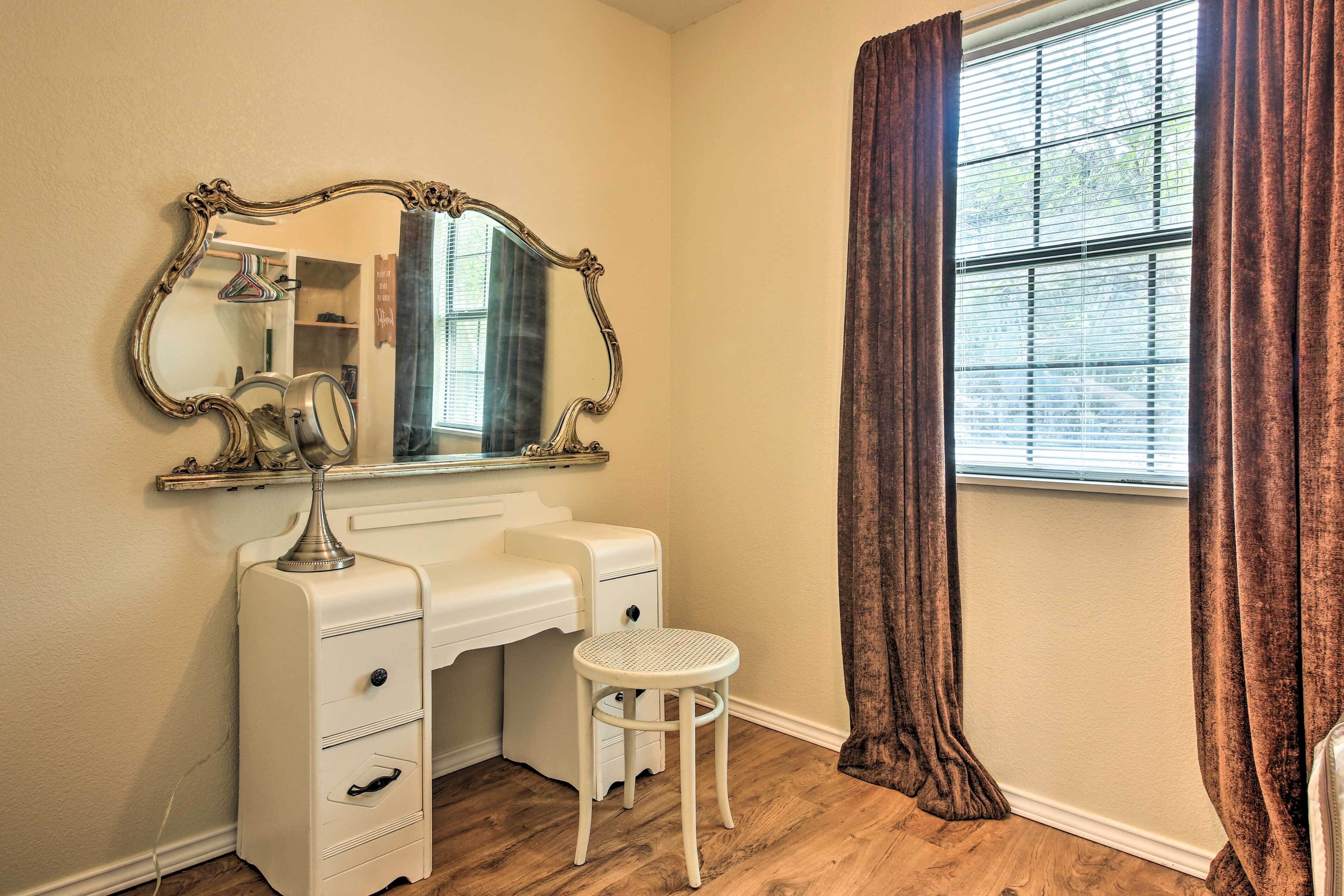 A room with a vanity, a mirror, a stool, and a window with curtains.