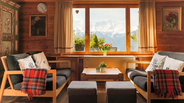 A cozy cabin interior with wooden walls, a large window with a mountain view, two sofas with plaid blankets, and a small table with a vase of flowers.