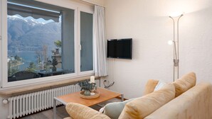 A room with a large window, a flat-screen TV mounted on the wall, a floor lamp, and a beige sofa. - Cosy apartment for 2 guests with WIFI, TV, balcony and pets allowed (Ascona)