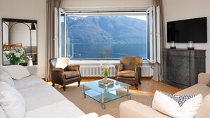 A living room with a large window offering a mountain and lake view, a sofa, two armchairs, a coffee table, a TV, and a cabinet. - Nice apartment for 4 people with WIFI, TV, balcony and pets allowed (Ascona)