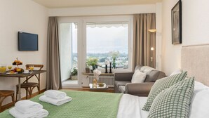 A hotel room with a bed, a sofa, a dining table with breakfast, a view of the city, and a window with curtains.