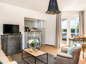 A modern living room with a flat-screen TV, a sofa, a coffee table, and a dining area with a table set for breakfast.
