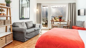 A modern hotel room with a grey sofa, a red bed, a dining area with a table and chairs, a large mirror, and a view of mountains through the sliding doors. - Lovely apartment for 2 people with WIFI, TV, balcony and pets allowed (Ascona)