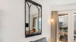 A living room with a grey sofa, a large mirror, and a view of a cityscape through a sliding glass door. - Lovely apartment for 2 people with WIFI, TV, balcony and pets allowed (Ascona)