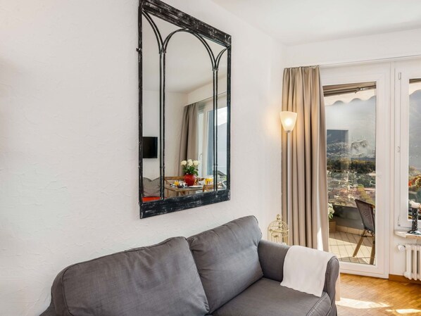 A living room with a grey sofa, a large mirror, and a view of a cityscape through a sliding glass door.