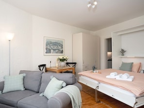 A modern bedroom with a grey sofa, a wooden dining table, a bed with rolled towels, a white wardrobe, and a painting on the wall.